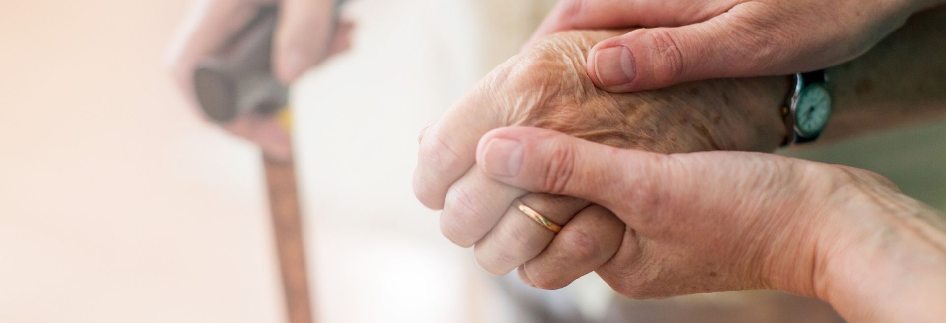 Eine jüngere Hand hält fürsorglich die faltige Hand einer älteren Person, die einen Gehstock hält. Symbol für Unterstützung und Fürsorge im Alter.